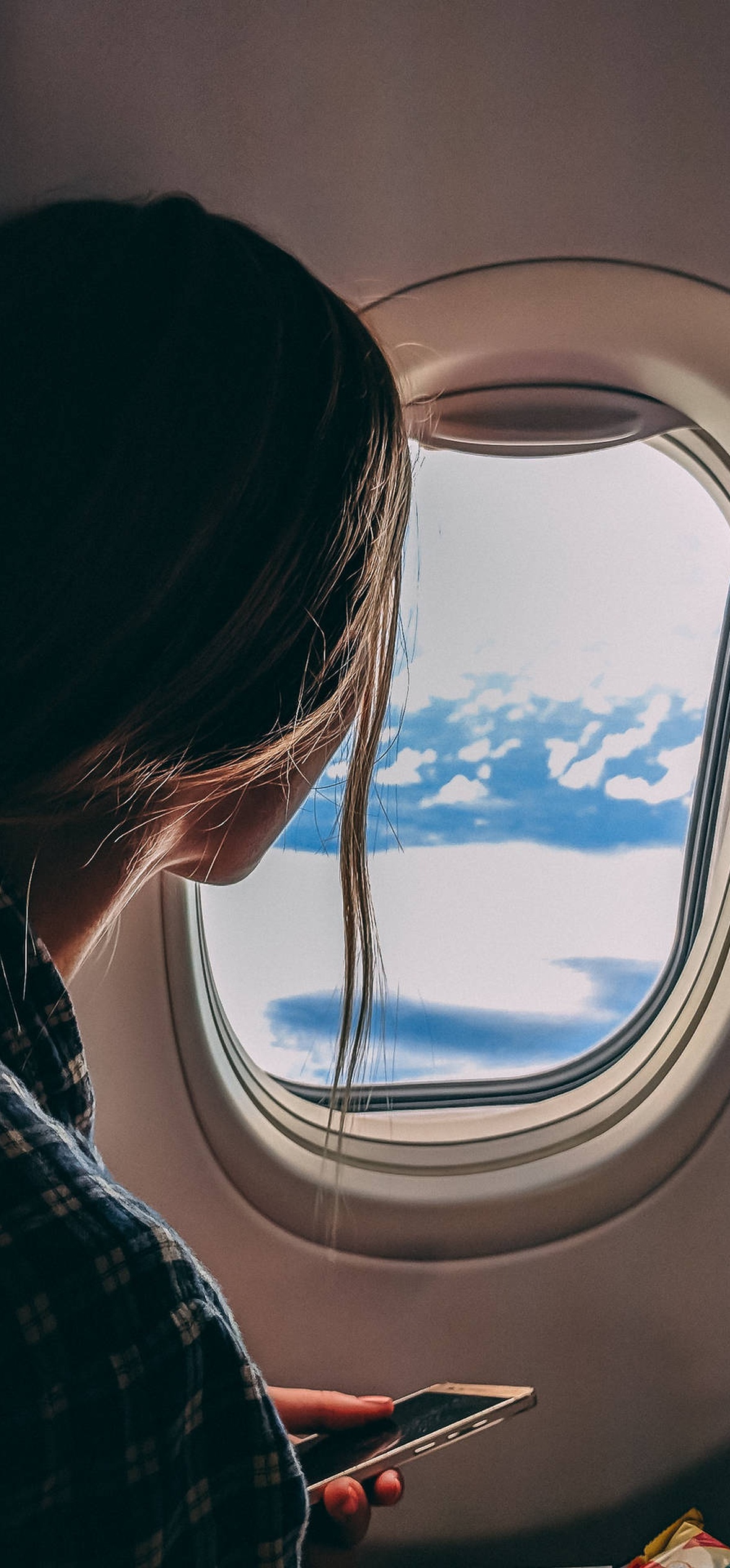 Person looking out airplane window at clouds while holding phone