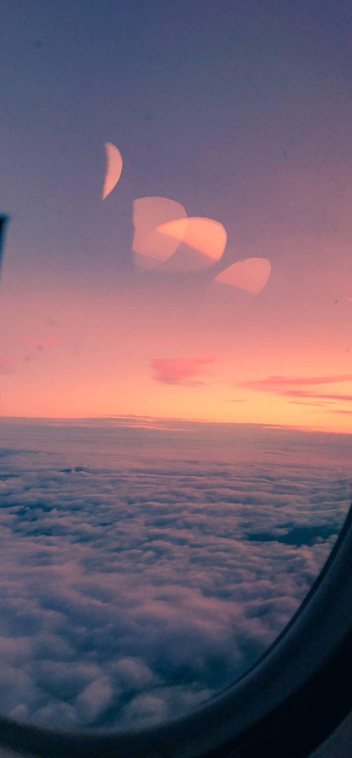 Aircraft window showing sunset over clouds with pink and purple hues and lens flare.
