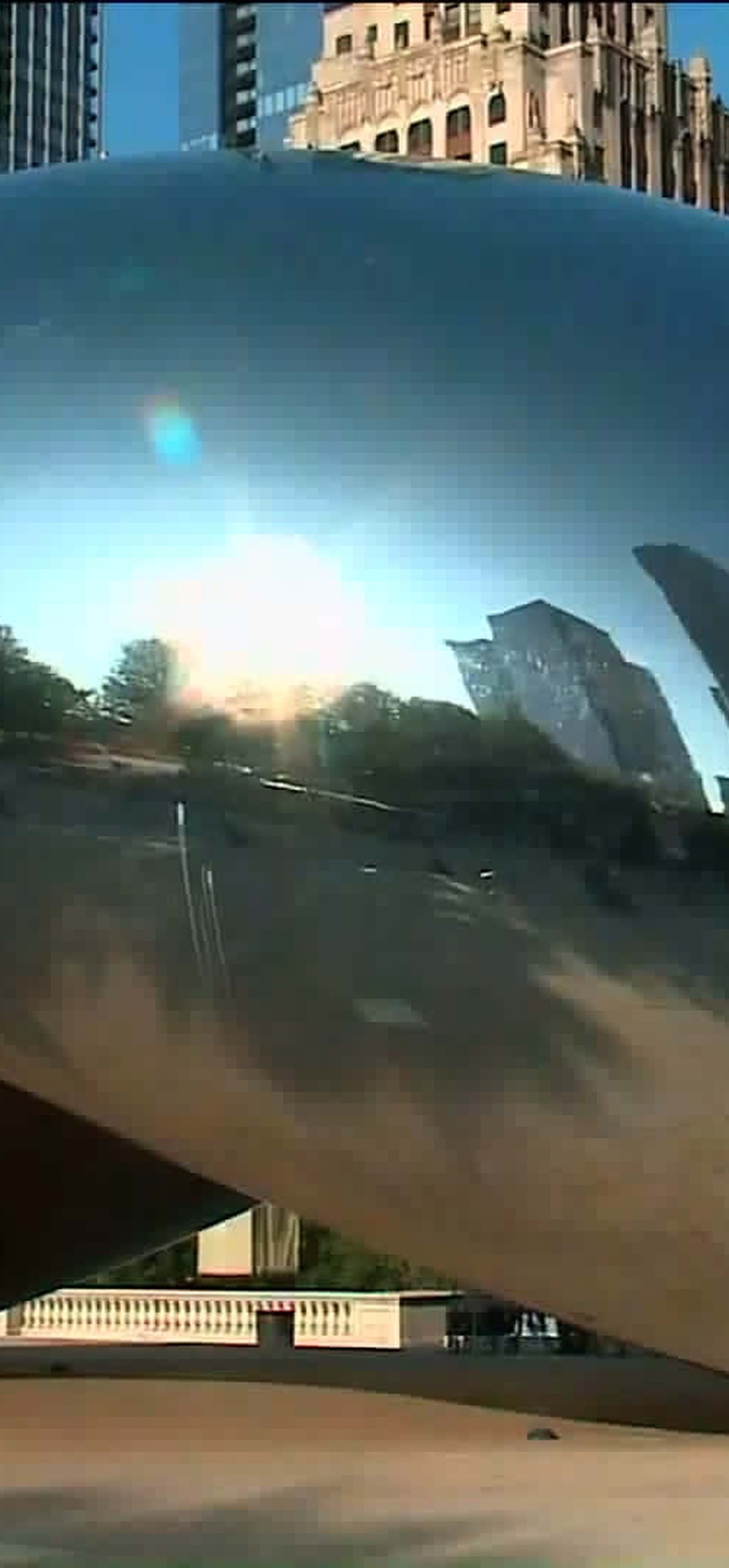 Shiny bean sculpture reflecting city buildings and trees at sunset.