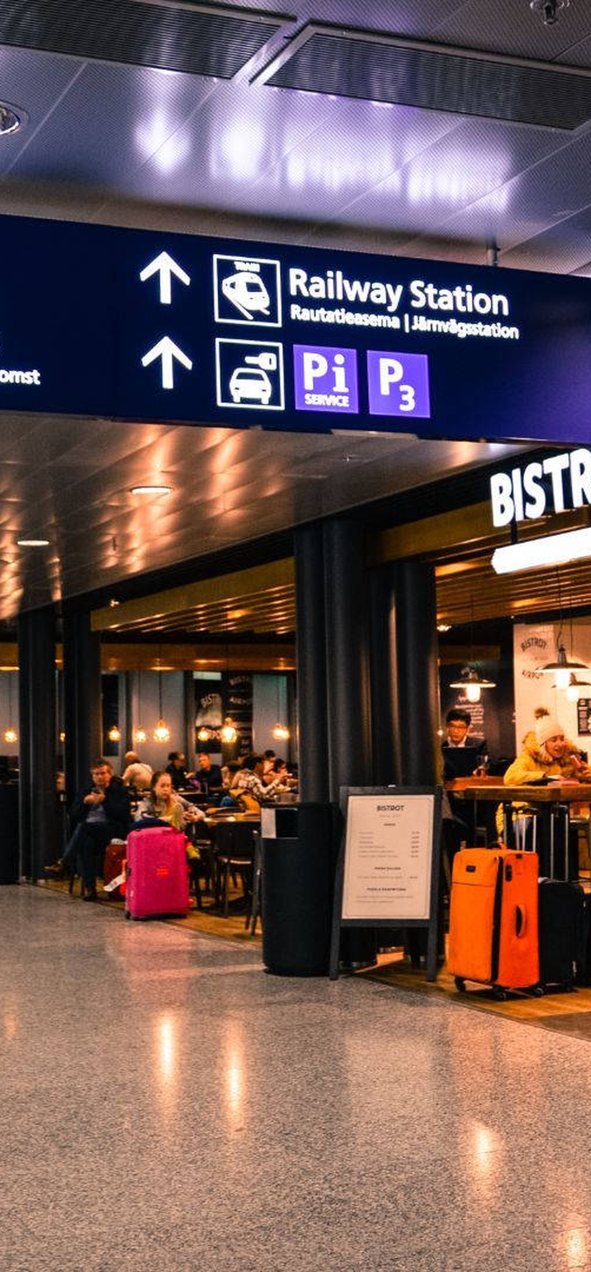 People with luggage walk through a busy airport terminal near a bistro.