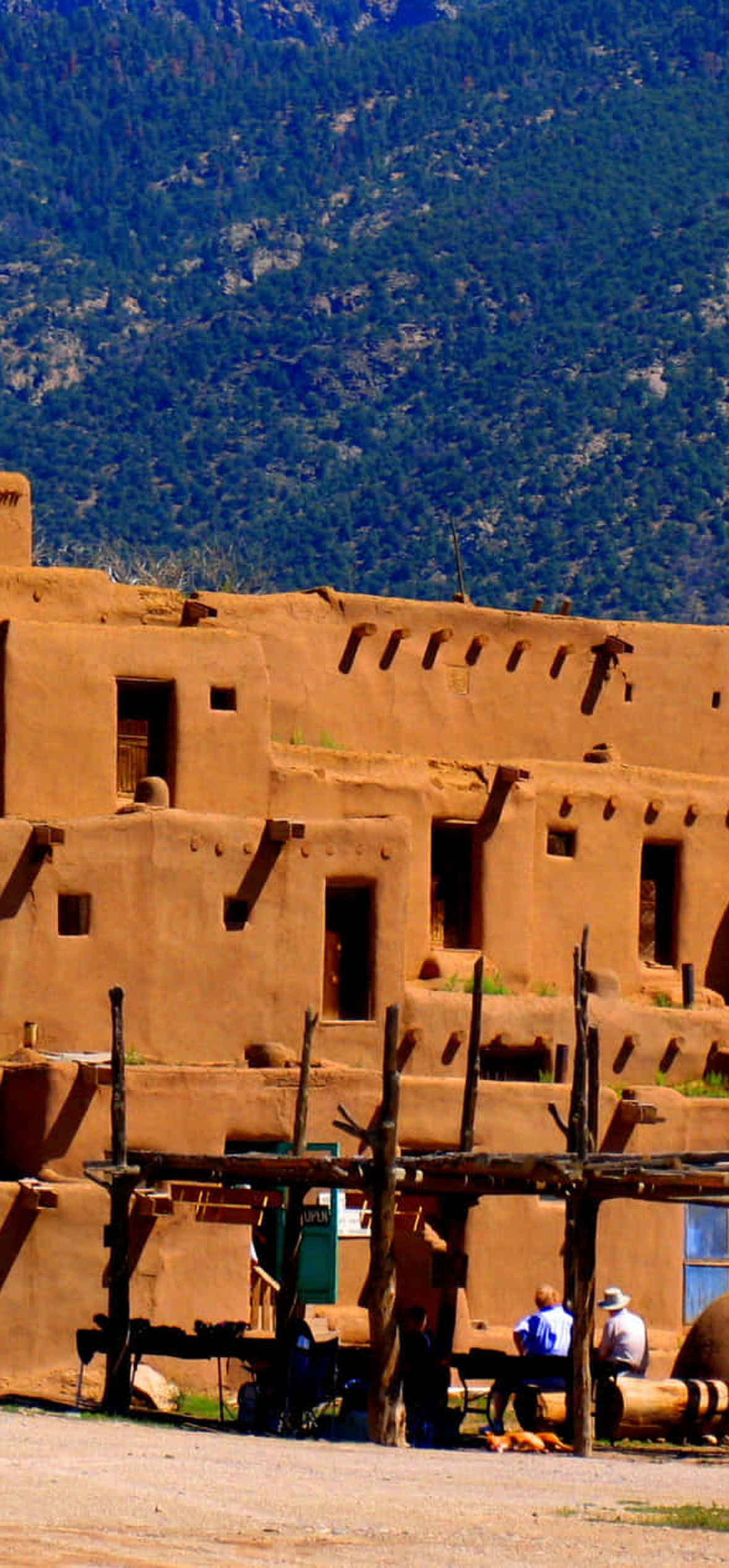 Adobe pueblo buildings with tourists near wooden structures against forested mountains.