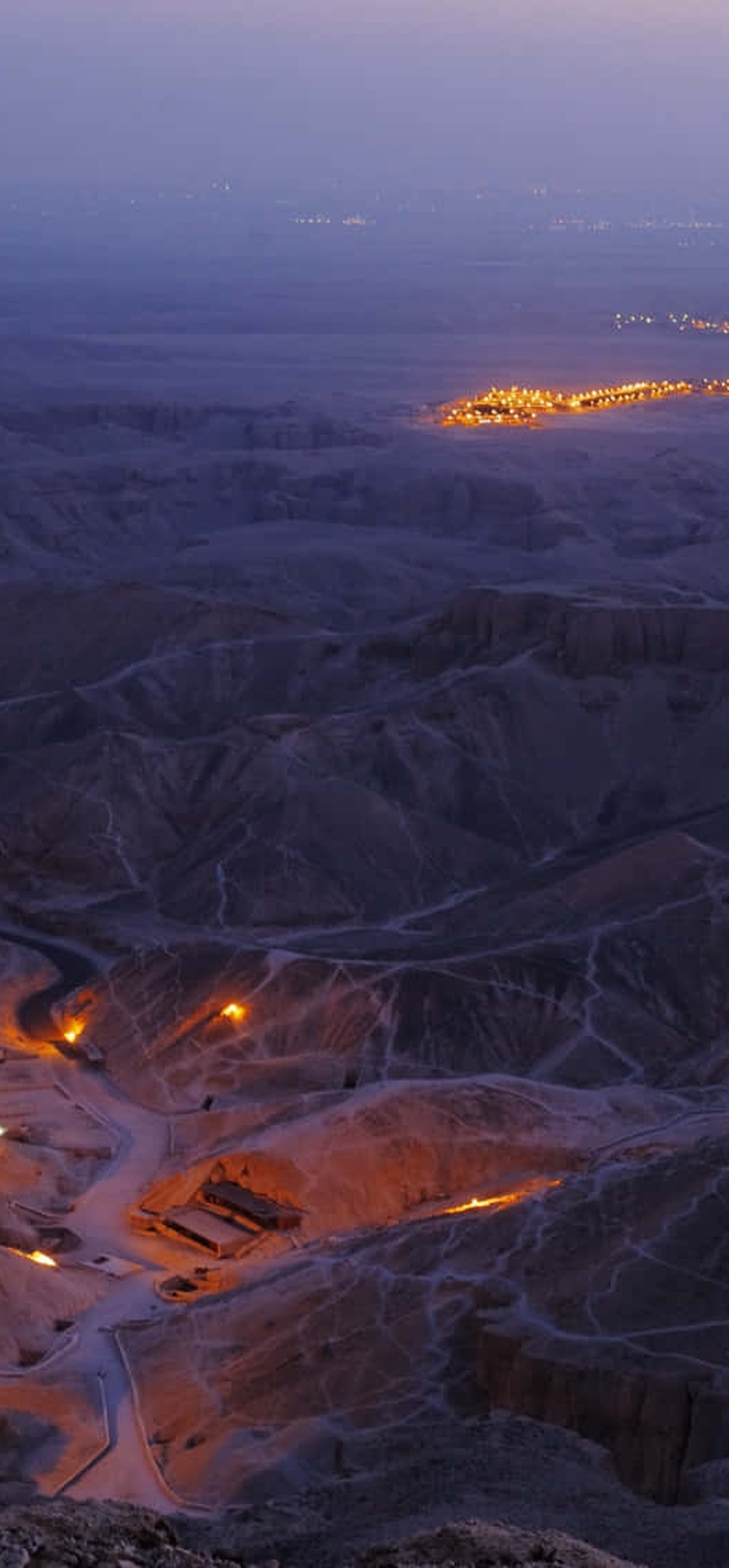 Desert canyon road lit by orange lights at twilight with distant city lights.